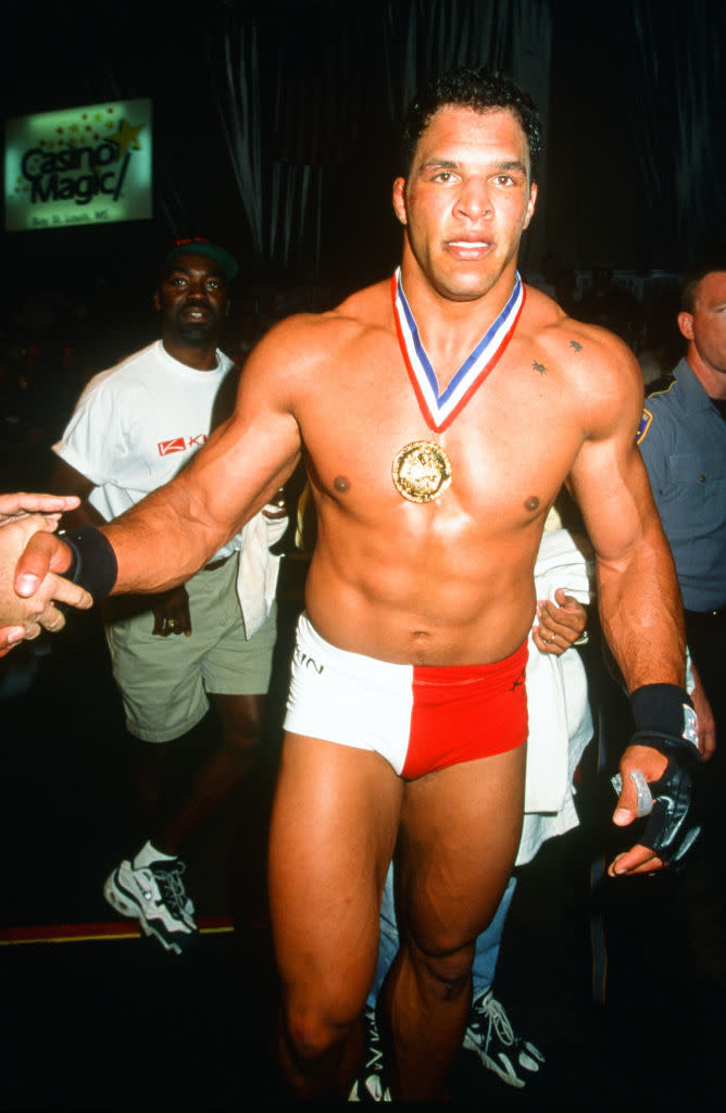 A muscular Kurt Angle wearing a medal and red and white trunks, walking on a red carpet, acknowledging the crowd