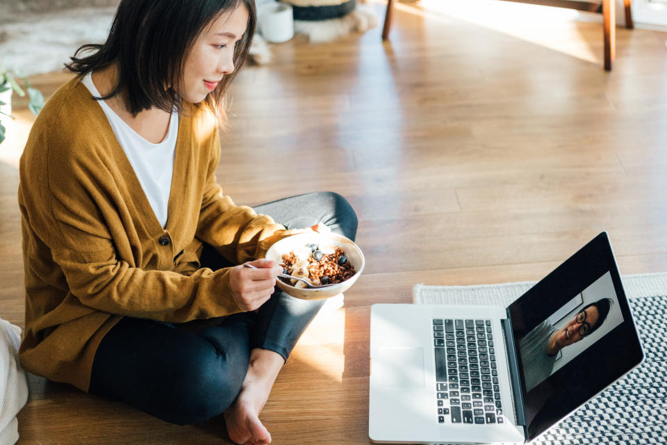 An Asian woman sits cross-legged on the floor as she eats a bowl of breakfast cereal while on a video call with an Asian man on her laptop