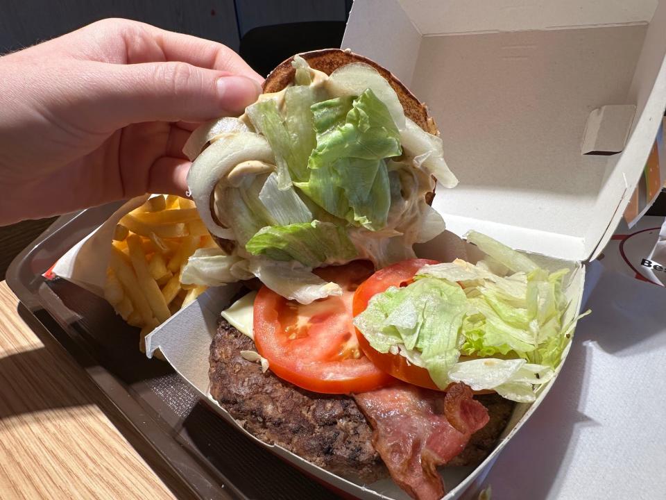 mcdonalds burger shiny with hand lifting the bun to show lettuce onion bacon tomato cheese on top of burger patty in white box next to french fries