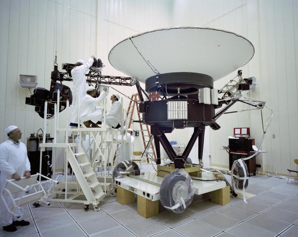 Several people in white coveralls work on the Voyager 2 spacecraft in a warehouse