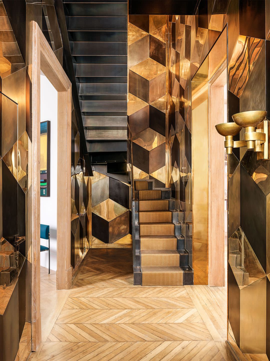 a stair hall has walls sheathed in polished and patinated brass plate in an interlocking cube pattern, two brass sconces, wood floor in a chevron pattern