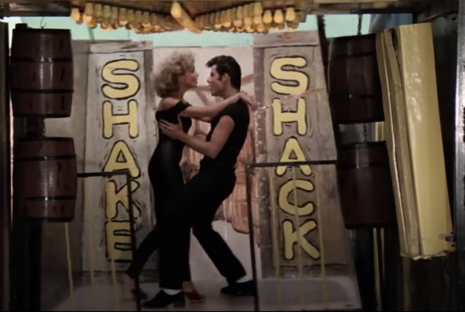 Olivia Newton-John and John Travolta dancing together in the movie between a &quot;Shake Shack&quot; sign doorway