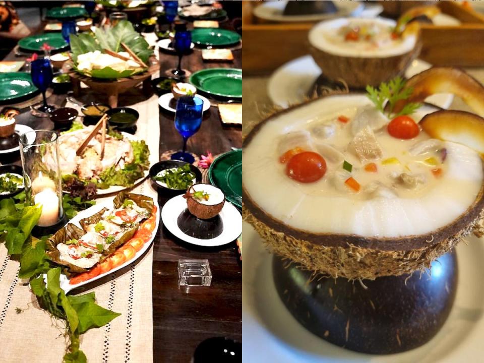 A table filled with fish, coconuts, and other plants with green plates and blue wine glasses for table settings; A coconut filled with a soup made with fish and vegetables