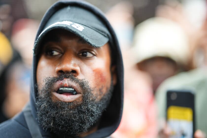 A man with a beard and mustache wears a baseball cap under a hoodie, plus a Balenciaga mouth guard