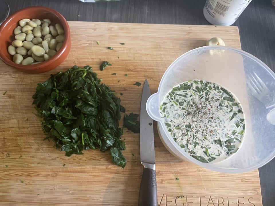 An overhead shot of a chopping board with a bowl of broad beans, a pile of chopped cooked spinach, and a jug with milk, cream, eggs and tarragon for the filling.