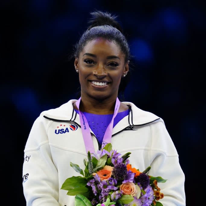 Simone Biles smiling with a medal, holding flowers and a plush toy at a gymnastics event