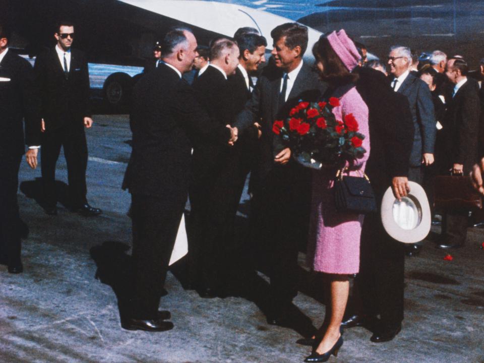 President John F. Kennedy and First Lady Jackie Kennedy wearing a bright pink skirt suit are greeted with flowers in front of Air Force One.