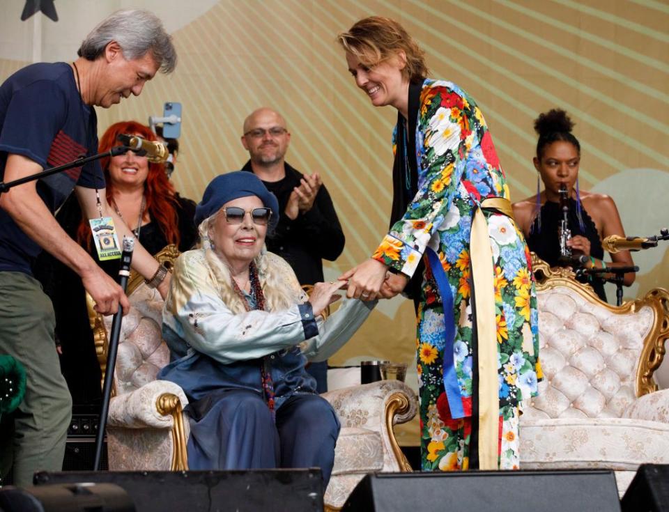 joni mitchell smiles while sitting in an armchair, she holds brandi carliles hands who stands to the right, mitchell wears a blue beret, large sunglasses, and a blue and white outfit, other people are also on the stage in the background