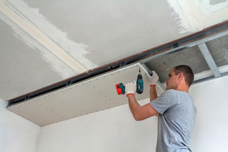 A worker in a grey shirt uses a drill to install a white ceiling. 