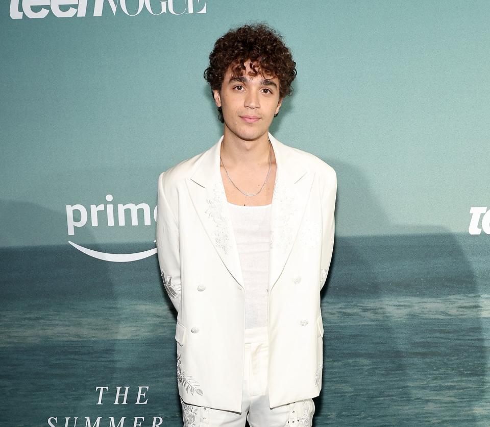 david iacono, a young man stands looking at the camera with a neutral facial expression, he has curly brown hair and wears a white tshirt, trousers and jacket