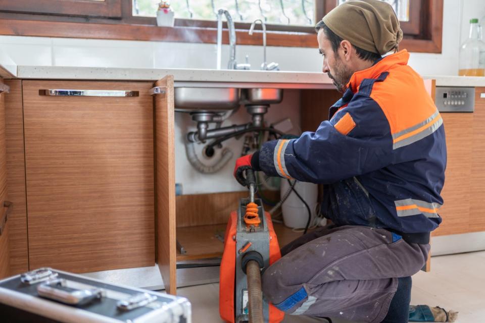 A plumber in a blue and orange jacket uses a tool to inspect under-the-sink plumbing. 