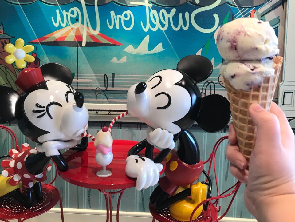 hand holding with cone full of gelato scoops in front of a gelato shop with mickey and minnie statue