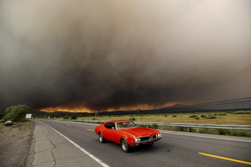 A car drives on a highway with the glow of flames and black smoke in the background.