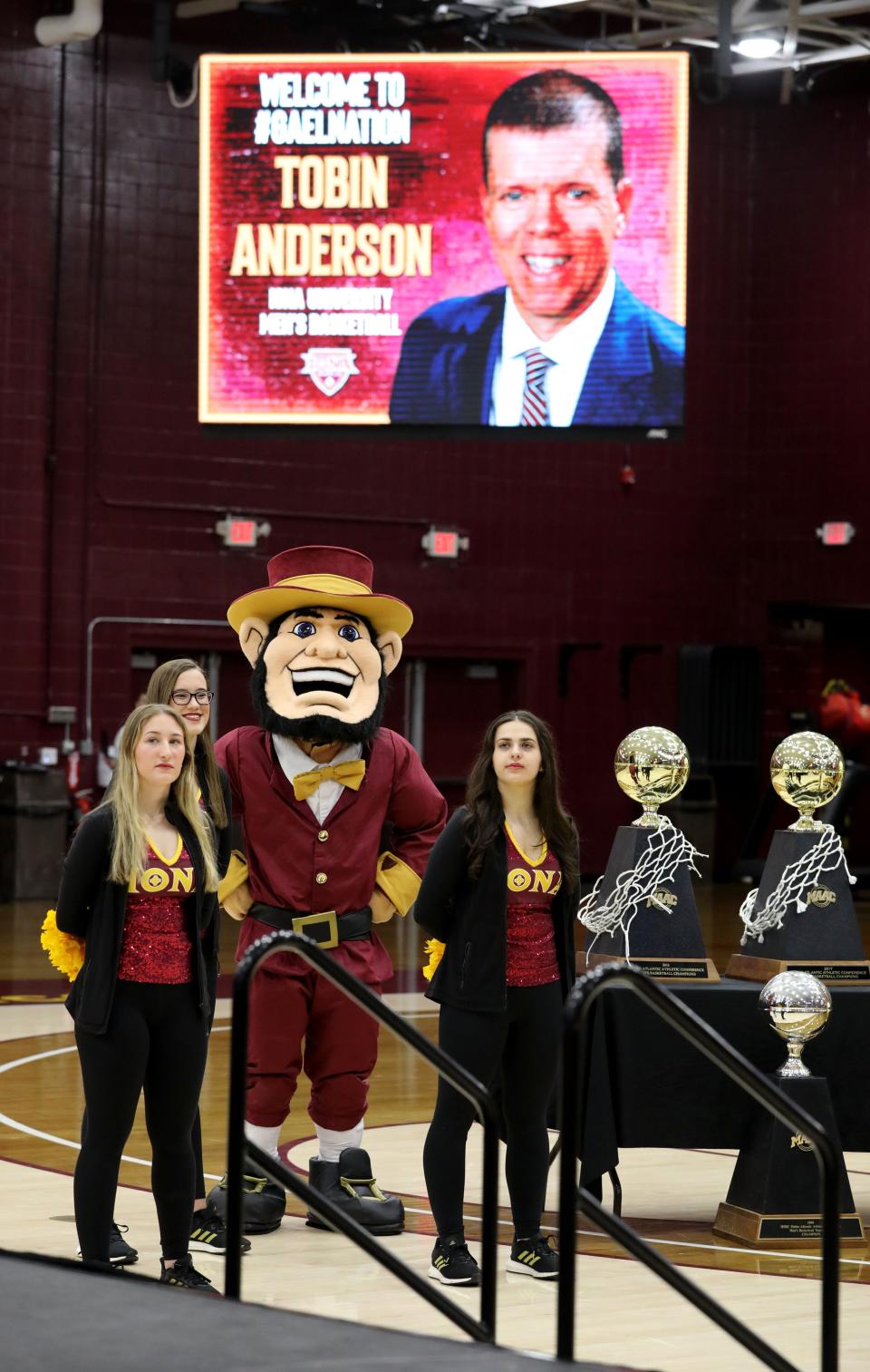 Iona University mascot and cheerleaders are pictured under a display board announcing Tobin Anderson as the new Iona University men's basketball coach, March 22, 2022.