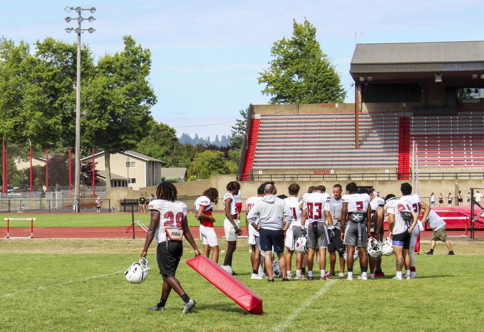 Players huddle with a member of the coaching staff during an NCAA college football practice, Wednesday, Aug. 9, 2023, in Monmouth, Ore. As one of just two NCAA Division II schools with football teams on the West Coast, Western Oregon spends hundreds of thousands of dollars in travel expenses to compete in the Lone Star Conference against schools in Texas and New Mexico. (AP Photo/Tim Booth)