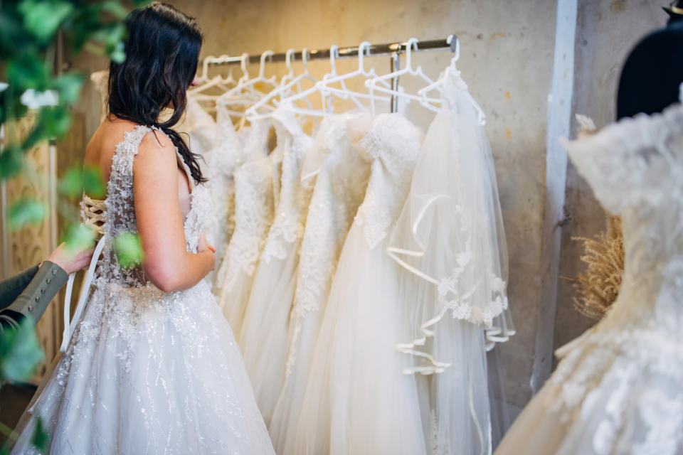 A bride-to-be, wearing a wedding dress, stands in front of a rack filled with various white wedding gowns, looking for the perfect one