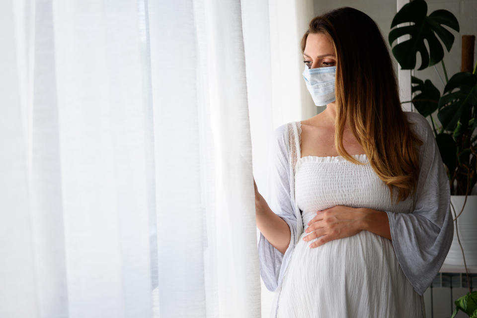 Pregnant woman standing by the big window with face medical mask on. Worries about child birth during pandemic.