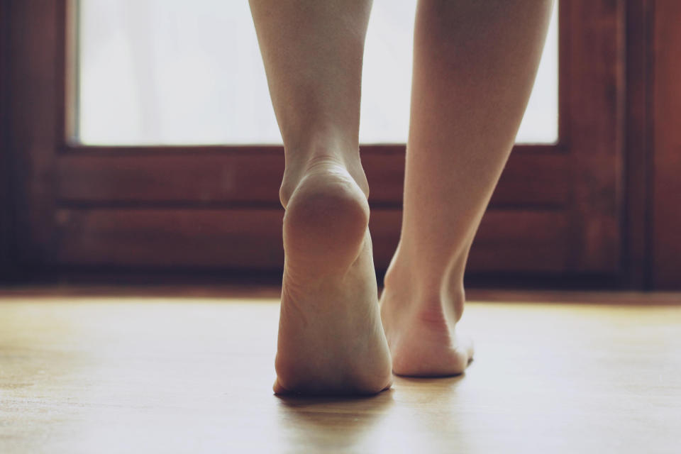 Close-up of a person's barefoot feet walking indoors on a wooden floor, with light coming from the window