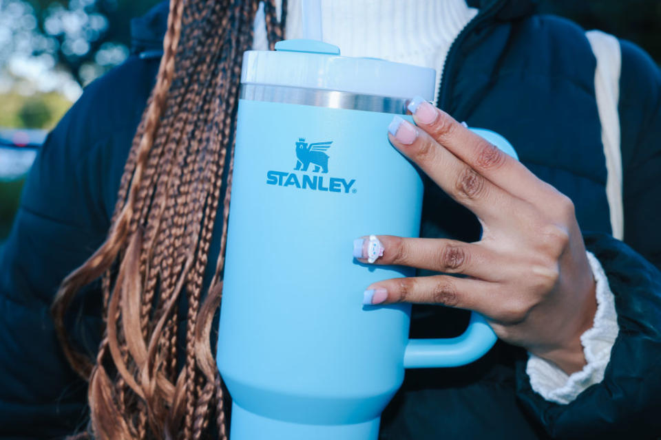 Person holding a large Stanley brand insulated travel mug with braided hair visible, wearing a jacket and a turtleneck sweater