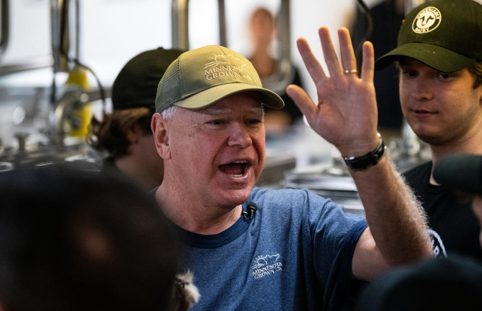 Man wearing a green cap and blue shirt gestures with his hand raised, surrounded by people in a kitchen setting