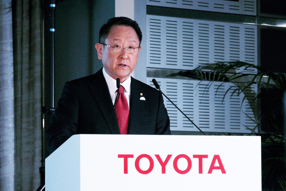 Toyota CEO Akio Toyoda, in a dark suit and red tie, speaking at a white podium with a red Toyota logo.