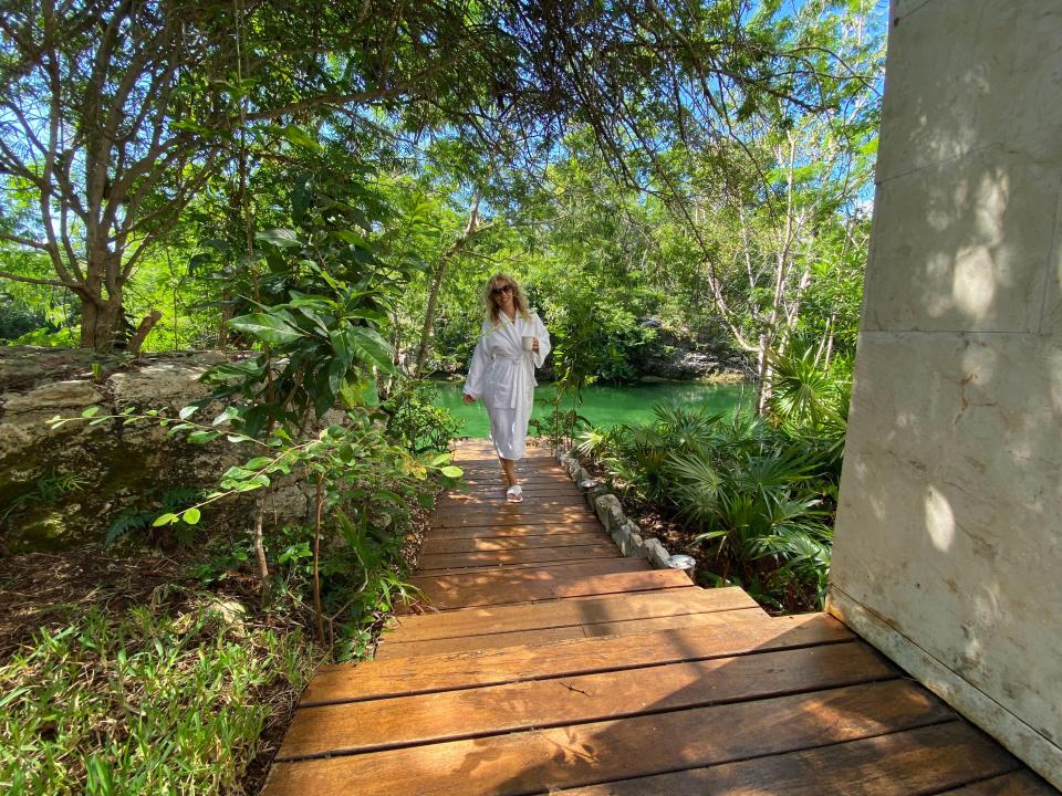 A blonde woman in a bathrobe posing next to a walkway framed by green trees.