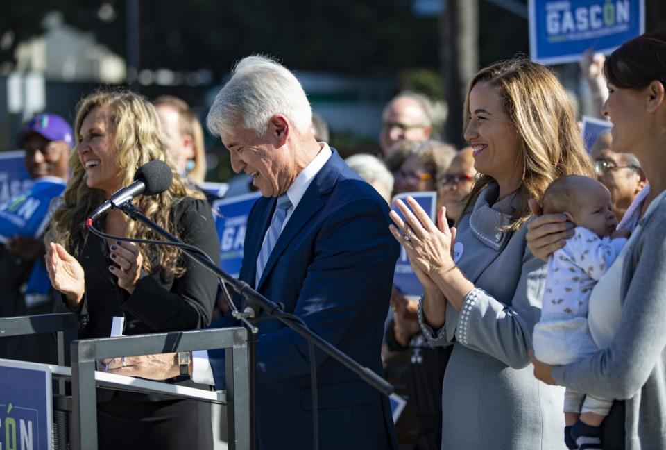 George Gascón, alongside three women, one holding an infant, smiles and speaks into a microphone.