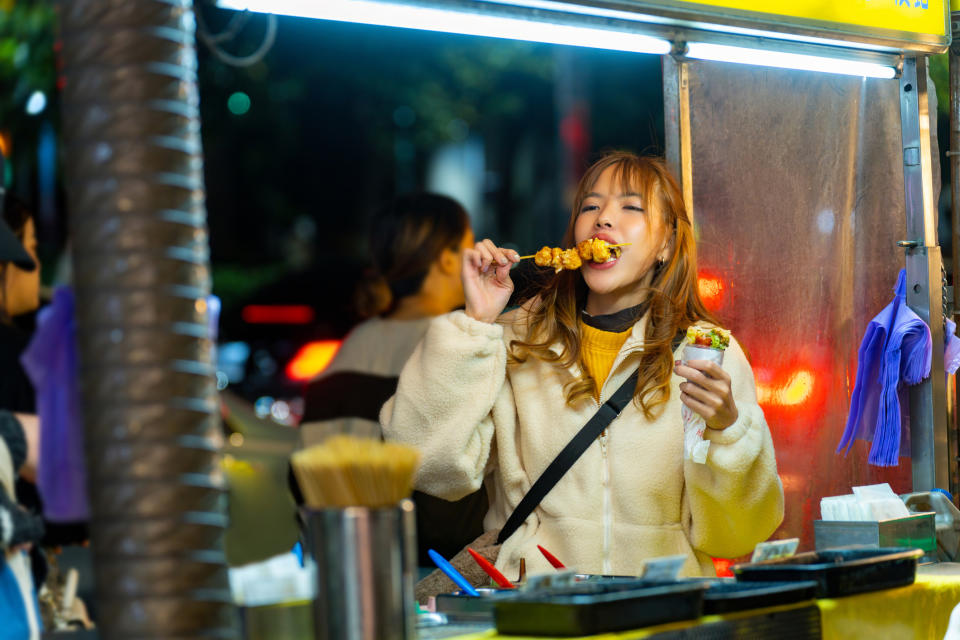 A person enjoys street food at a night market, biting into a skewer and holding a cup with other snacks. There are various foods and utensils on the counter.