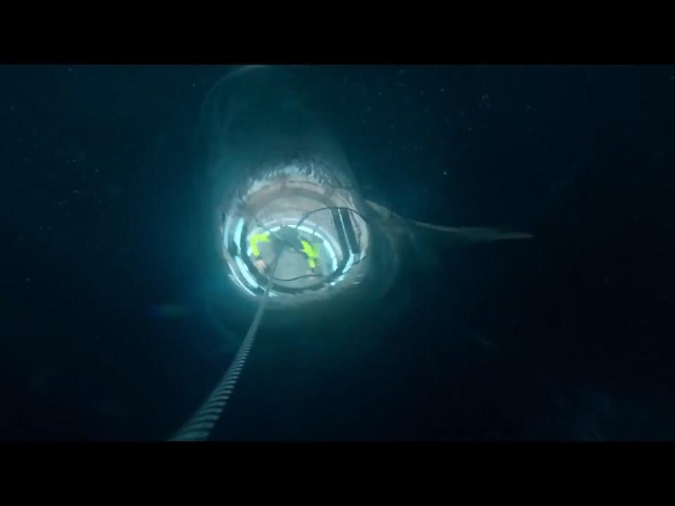 A still from "The Meg" shows the megalodon seen from above, hoding the shark cage in its mouth and drawing it down to the abyss.
