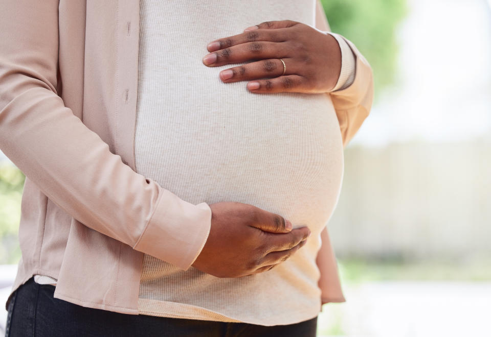 Pregnant person cradling their belly with both hands, wearing a beige top and black pants