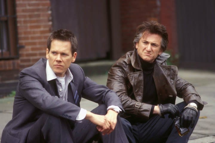 Kevin Bacon and Sean Penn sit on a sidewalk next to each other.