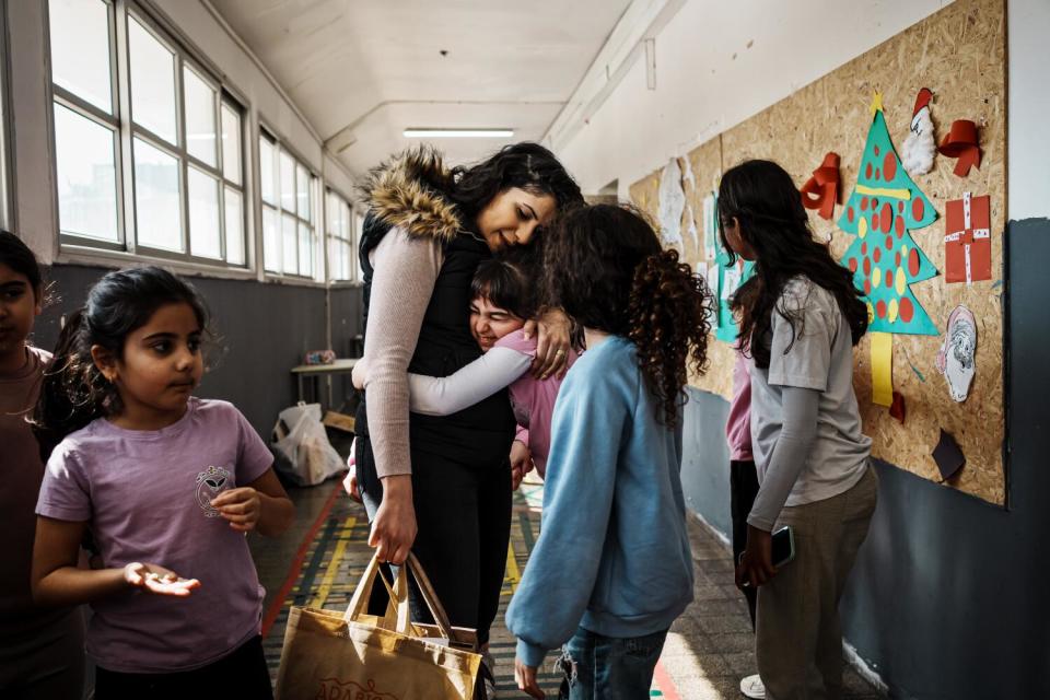 A woman with dark hair, holding a bag, gets a hug from a girl as others stand nearby