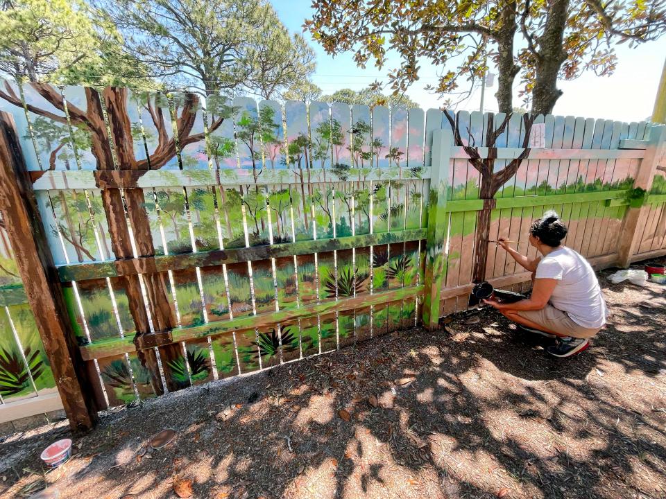 Arts and Design Society member Robin Chastain works on a mural of the Florida National Scenic Trail along the backyard fence at the Emerald Coast Science Center. The mural is part of a fundraiser the arts group is holding to help cover costs of programs and building repairs.