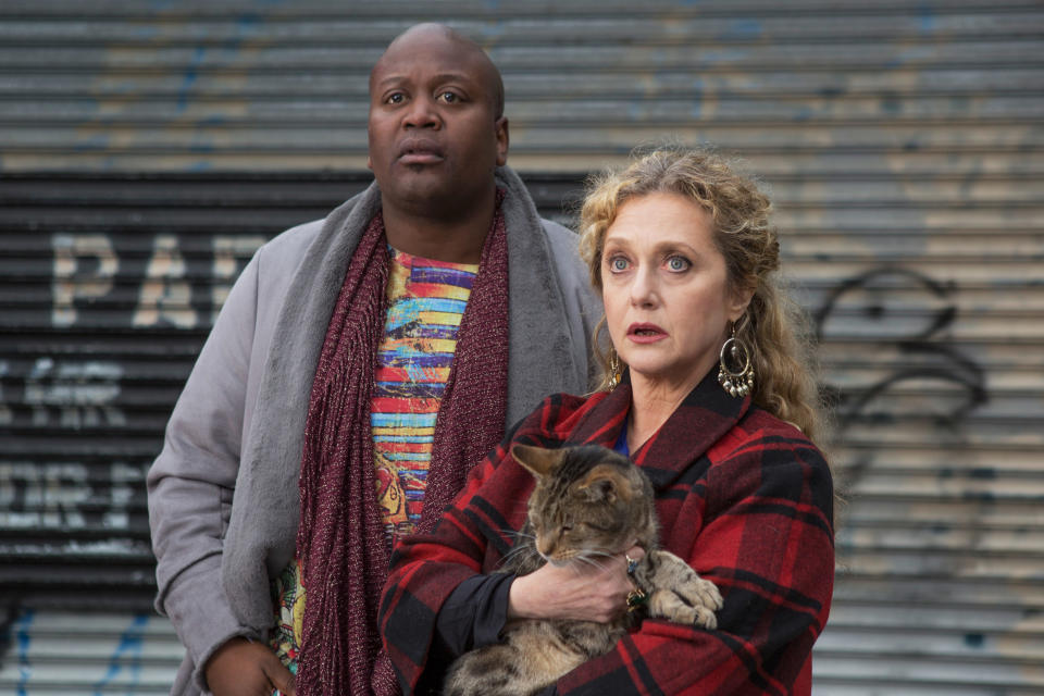 Two actors from the TV show 'Kimmy Schmidt' posing with a cat, man in scarf and woman in plaid coat