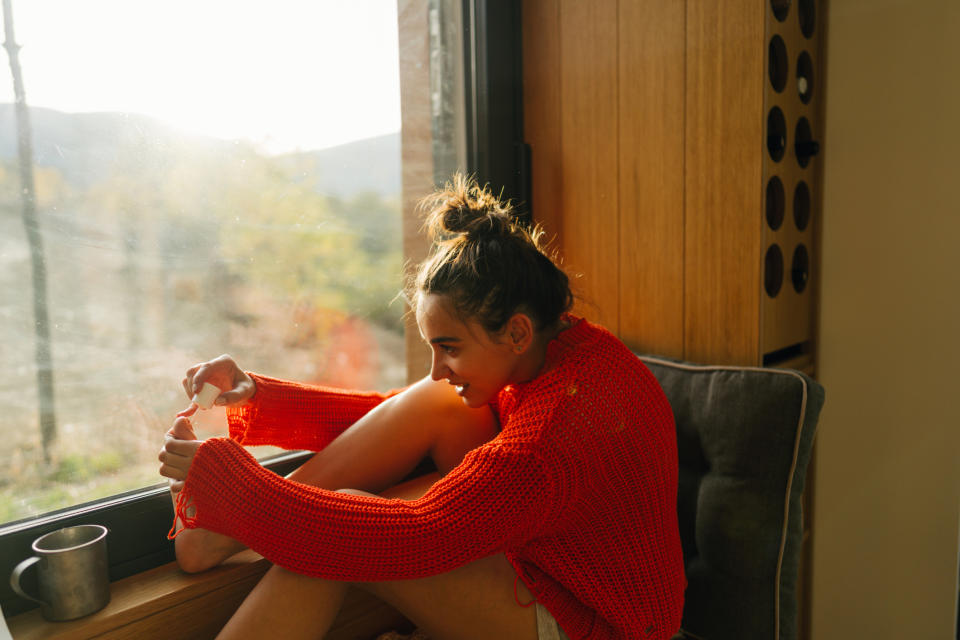 Woman seated by window, smiling, in cozy red knitwear, applying toenail polish