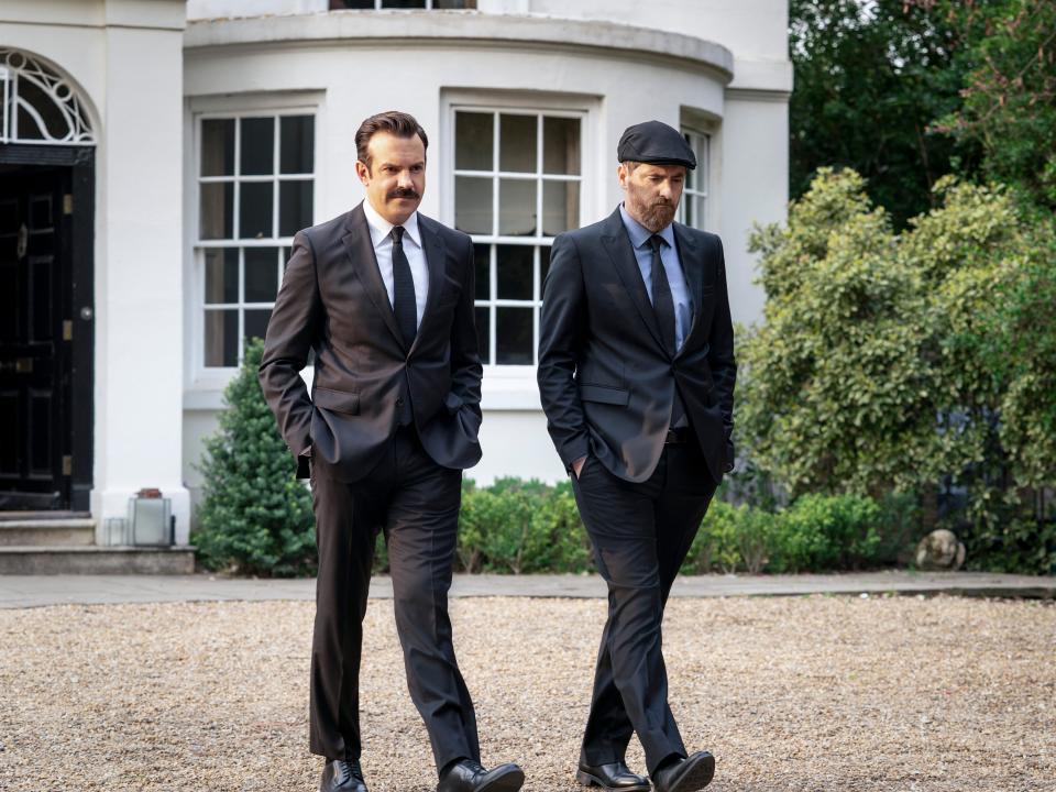 jason sudeikis and brendan hunt in ted lasso wearing suits and walking on gravel away from a white building