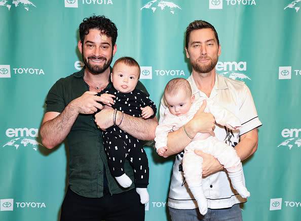 Michael Turchin and Lance Bass hold their twin babies at an EMA event. They are standing in front of a banner with Toyota and EMA logos