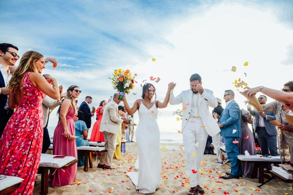 A man and woman exit their beach wedding as people throw flower petals over them.
