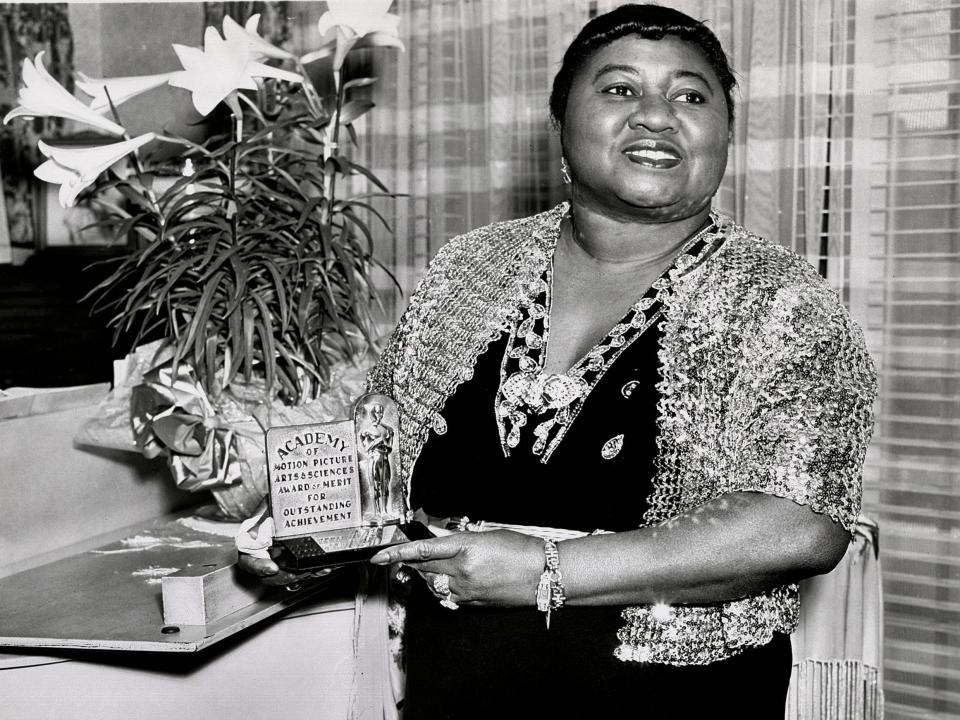 Black and white photo of Hattie McDaniel in a dress smiling and holding her award plaque.