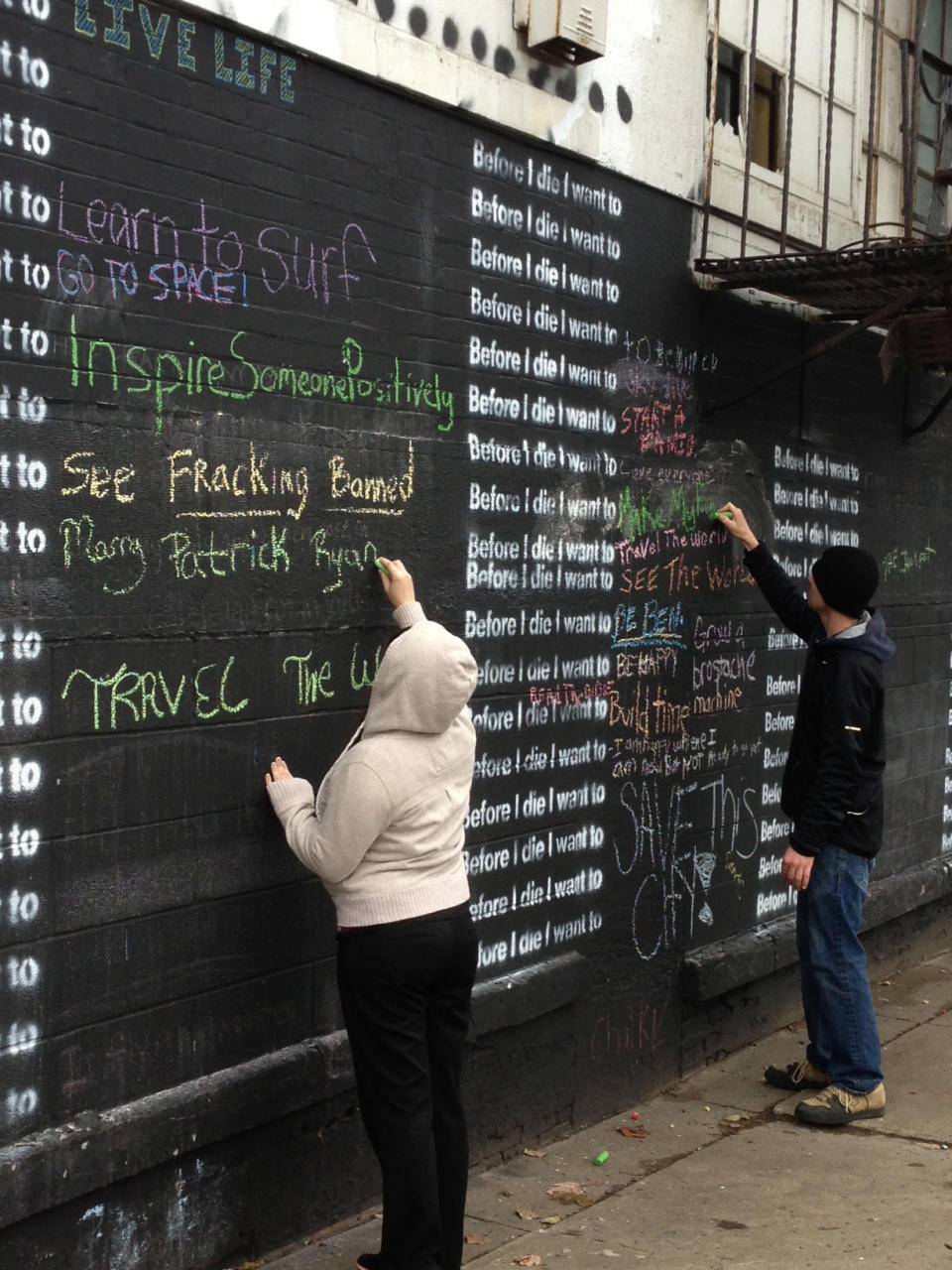 In this Nov. 7, 2013 photo, Sara McAlister, 22, and Patrick Kraushaar, 26, write on a wall in Syracuse, N.Y., that invites passers-by to complete the sentence: “Before I die, I want to...” In a phenomenon spreading across the globe, the oversize blackboards are being painted on buildings and freestanding displays. Since artist Candy Chang created the first wall on an abandoned house in her New Orleans neighborhood in 2011, more than 400 walls have gone up in the United States and more than 60 other countries. (AP Photo/Carolyn Thompson)