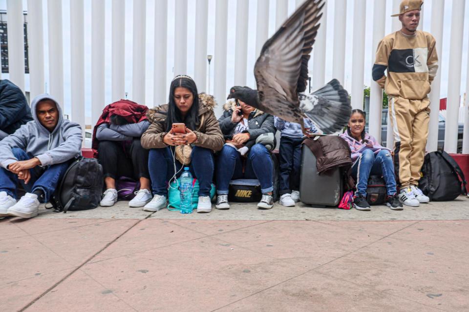 A pigeon flies near a row of people seated on the ground with their luggage