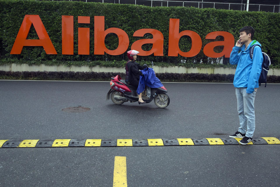 FILE - In this May 27, 2016, file photo, a man talks on his phone as a woman rides on an electric bike past a company logo at the Alibaba Group headquarters in Hangzhou in eastern China's Zhejiang province. China’s market regulator on Monday, Dec. 14, 2020 said it fined Alibaba Group and a Tencent Holdings-backed company for failing to seek approval before proceeding with some acquisitions. (AP Photo/Ng Han Guan, File)