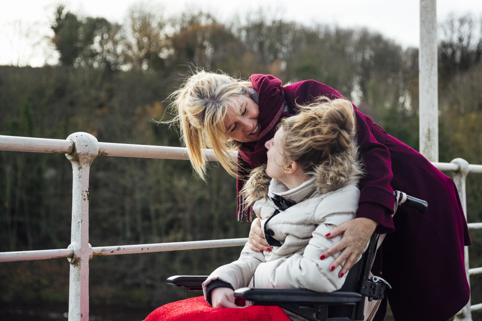 A mature caucasian woman and her teenage daughter who is using a wheelchair and has physical and mental disabilities. They are outdoors, on a bridge and they are face to face sharing a lovely moment.