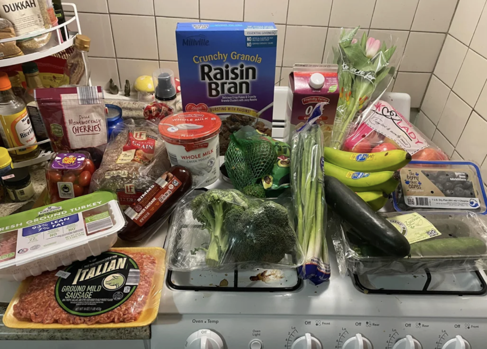 A variety of groceries placed on a stove and countertop, including fruits, vegetables, cereals, and meats, suggesting a recent shopping trip