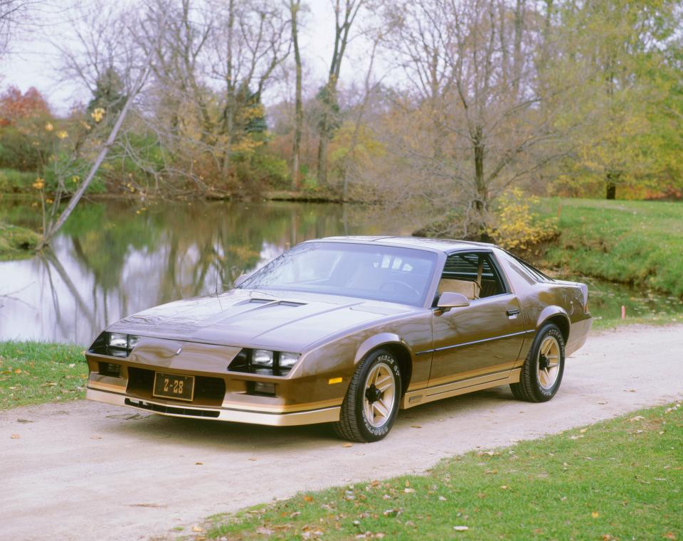 A brown 1982 Chevrolet Camaro Z28 parked on a driveway, with grass and trees surrounding it.