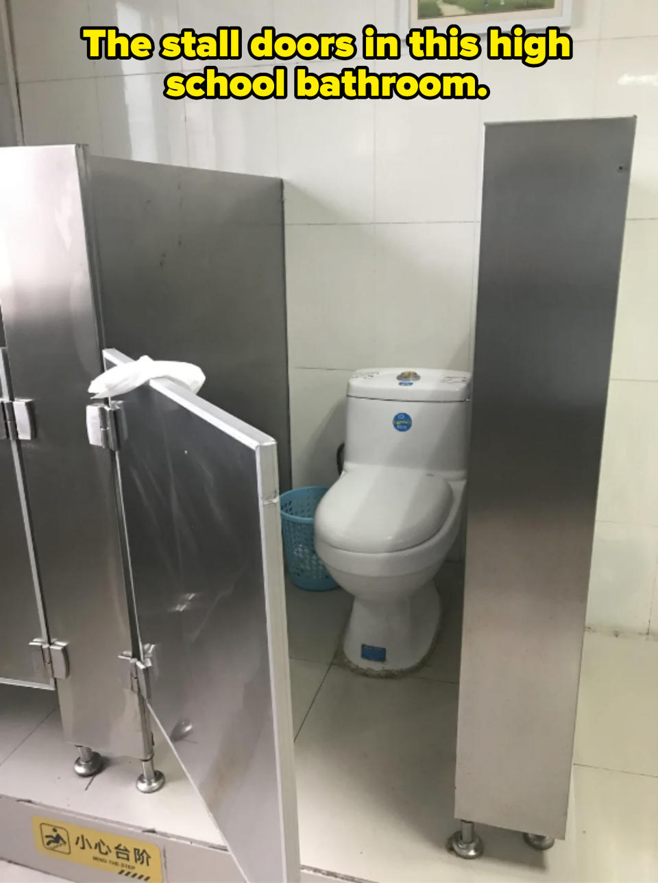 Bathroom stall with an open stainless steel door revealing a toilet and a blue wastebasket. There is a caution sign on the floor