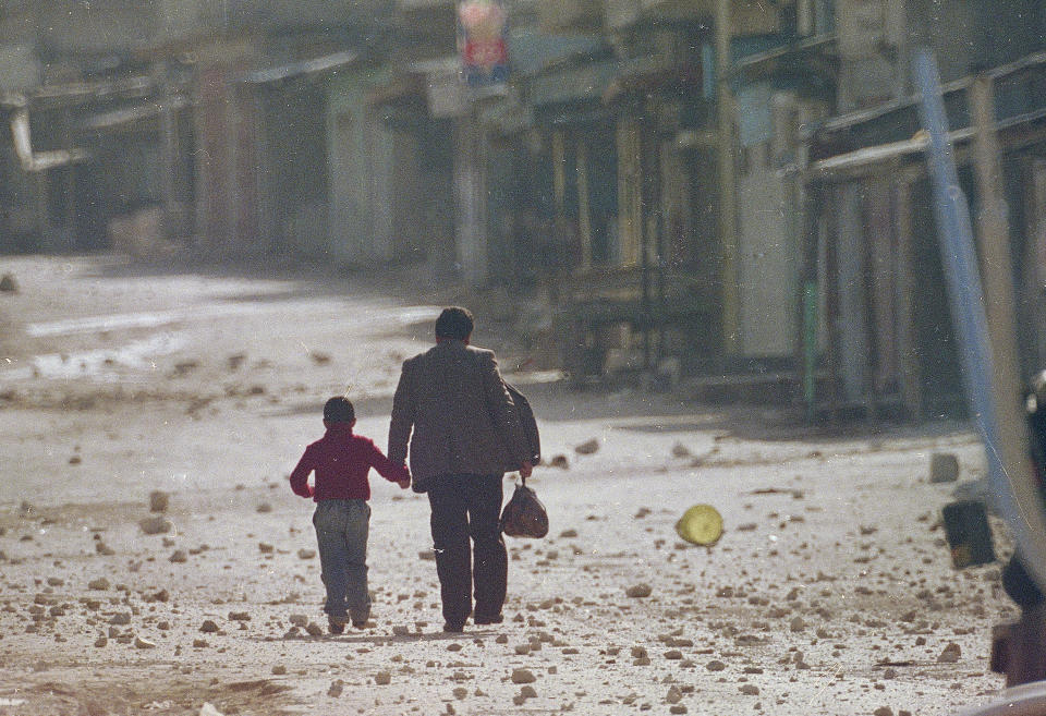 FILE - In this Dec. 11, 1987 file photo, a man holds his son's hand as they make their way along a debris littered street in the Balata refugee camp near Nablus in the occupied West Bank after a curfew was imposed on the camp. Western countries are reeling from the coronavirus pandemic, awakening to a new reality of economic collapse, overwhelmed hospitals and home confinement. But for millions across the Middle East and in conflict zones elsewhere, much of this is familiar. (AP Photo/Max Nash, File)