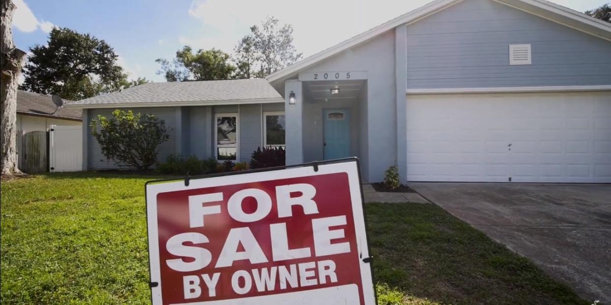 A photo of a blue house with a red "For sale by owner" sign in the front yard.