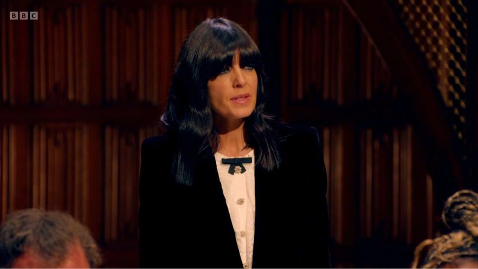 claudia winkleman in the traitors series 2 episode 4, wearing a very small bow, a white shirt and a black jacket, with her eyes barely visible beneath her fringe