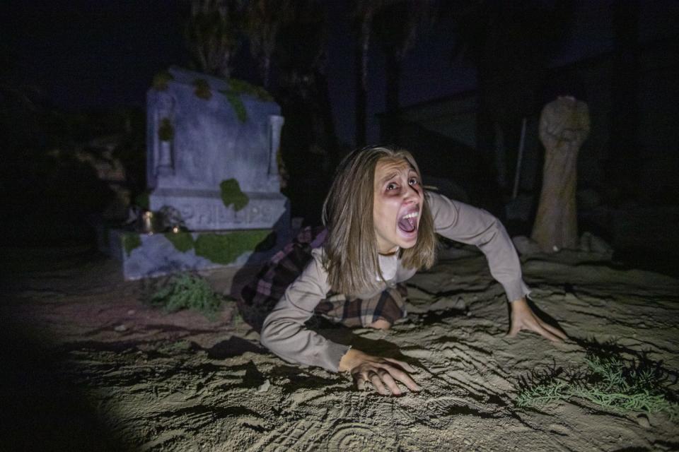 A woman with her mouth open kneels in the dirt in front of a graveyard scene